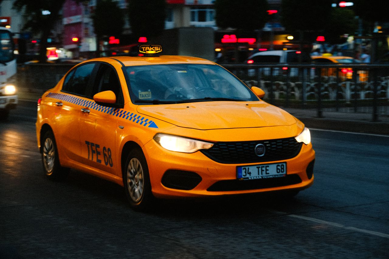 who-we-are Yellow taxi driving on a busy city street at dusk with blurred lights in the background.