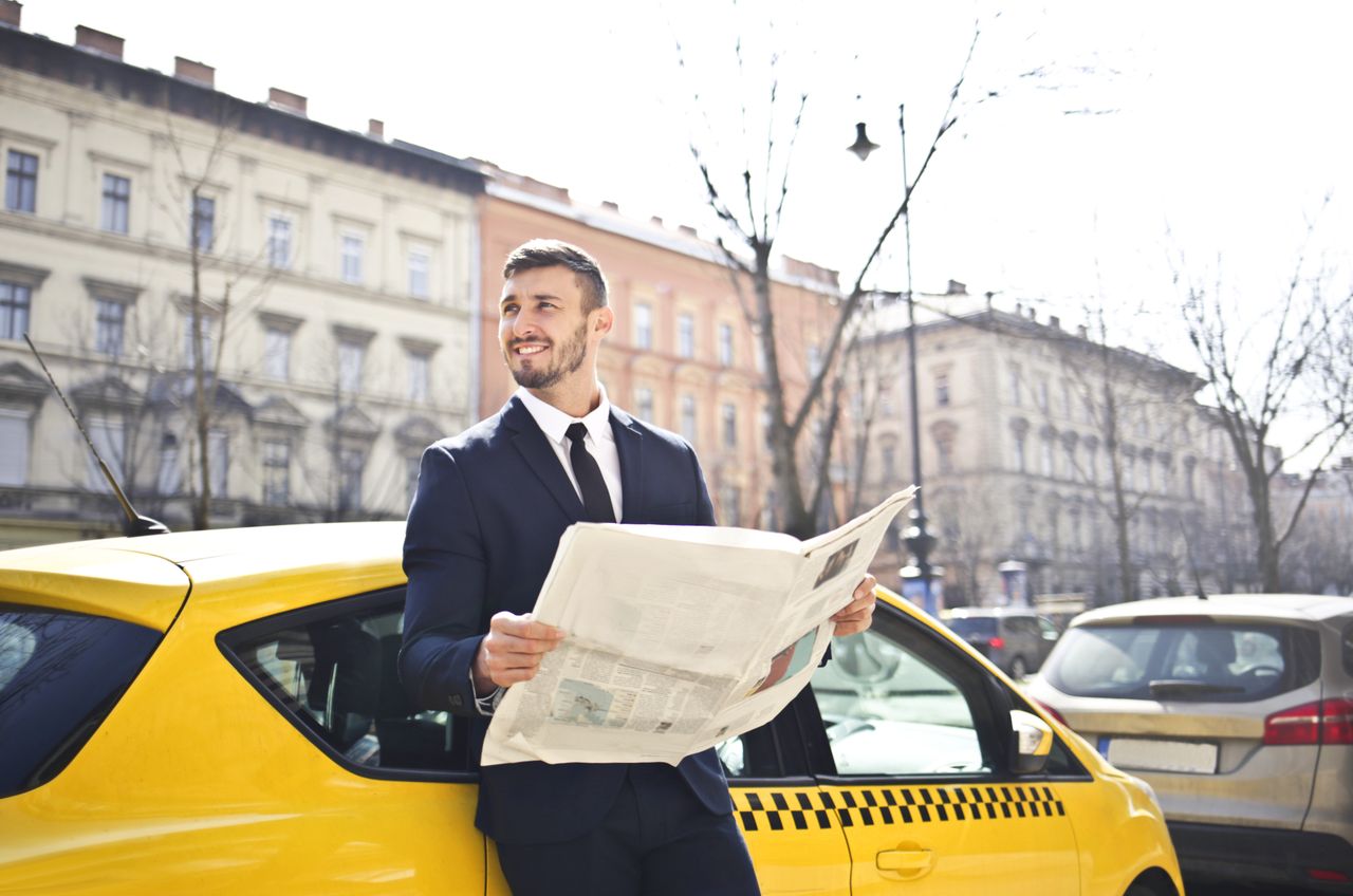 why-choose-us A businessman in a suit reads a newspaper while leaning on a yellow taxi in a city street.