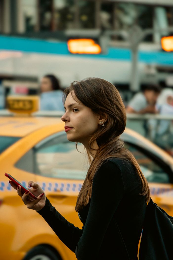 Young woman in city setting using smartphone near a yellow taxi cab.