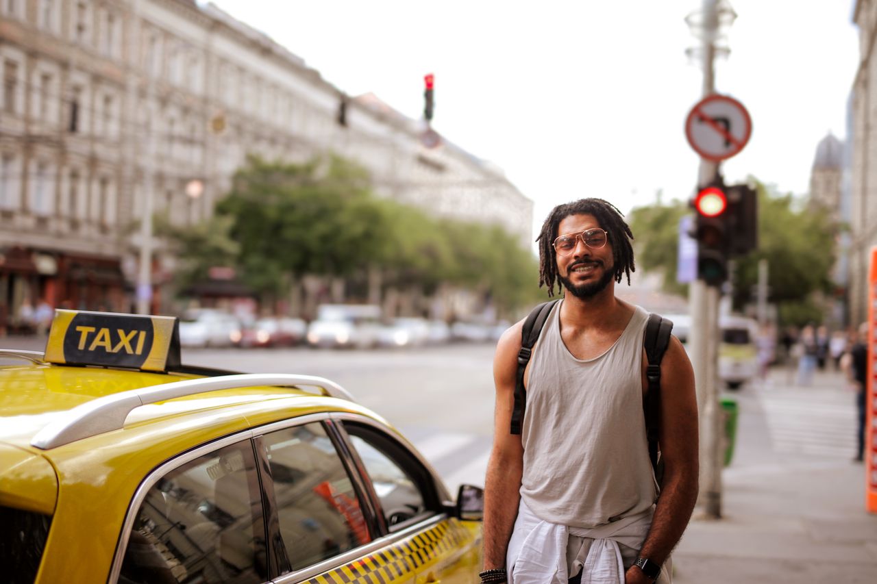 services-01 Smiling man beside a yellow taxi on a busy city street during the day.